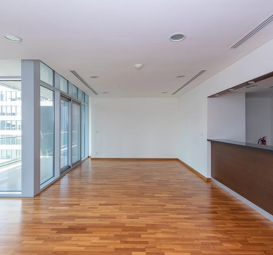 Large, empty living space with wood flooring, full-length windows, and a built-in counter or bar area.
