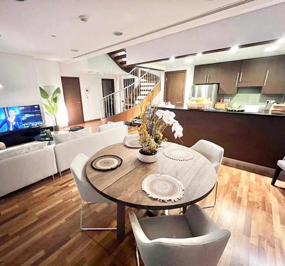 Open-plan dining space with a round wooden table, white chairs, and a sectional sofa facing a television.